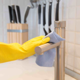 Mão com luva amarela a desinfetar torneira de cozinha, com um pano azul